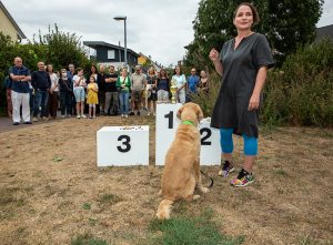 Eine Frau steht mit ihrem Hund vor einem Siegerpodest. Auf den verschiedenen Siegertreppchen sind Snacks für den Hund platziert. Auf dem Treppchen für den ersten Platz steht außerdem ein Pokal.