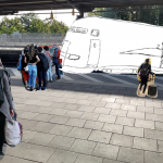 Ein Bahnhof mit einem leeren Bahnsteig. Über ein Bildbearbeitungsprogramm wurden Gruppen von wartenden Reisenden eingefügt. Auf dem Gleis wurde die Zeichnung eines Zuges eingefügt.
