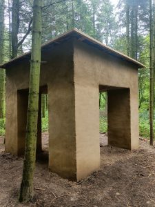 Eine Skulptur im Wald. Es ist eine Hütte aus Lehm. Sie öffnet sich in vier Türen.