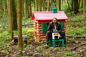 Jan Philip Scheibe sitzt vor einer Spielzeughütte im Wald. Vor ihm steht ein Schemel mit einem Kaffekocher.