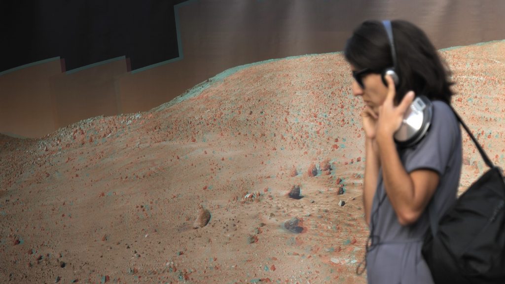 Eine Frau steht mit Kopfhörern vor einer Großbildablichtung einer Marslandschaft.