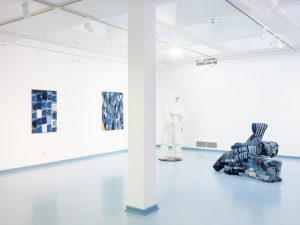 Ein Ausstellungsraum mit Plastiken und Gemälden. Die Gemälde bestehen aus geometrischen Figuren in Blautönen. Die Plastiken sind aus verschiedenen Stoffen.