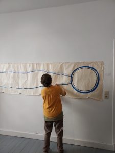Die Künstlerin Eca Wal steht zu einer Wand gedreht. Dort hängt eine Papierbahn. Zwei blaue Linien verbinden sich zu einem Kreis. Eva Wal beschriftet eine Linie.