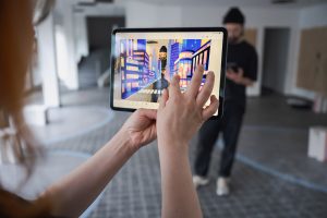 Eine Künstlerin hält ein Tablet vor sich. Auf dem Tablet ist der ihr gegenüberstehend Künstler zu sehen, wie er in einer virtuellen Stadt steht.