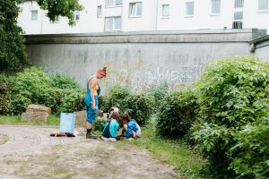 Zwei Kinder bemalen einen Mülleimer. Anne Brauer steht ihnen zur Seite.