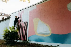 Eine Person besprüht die Wand mit Farbe. Das Mural hat Muster in den Farben Hellrot, Gelb, Blau und Schwarz.