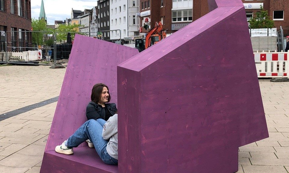 Inmitten eines Platzes in der Innenstadt steht eine Skultpur. Sie ist kantig, hat viele Öffnungen und ist violett. Menschen interagieren mit der Skulptur, indem sie darauf sitzen, liegen oder darunter stehen.