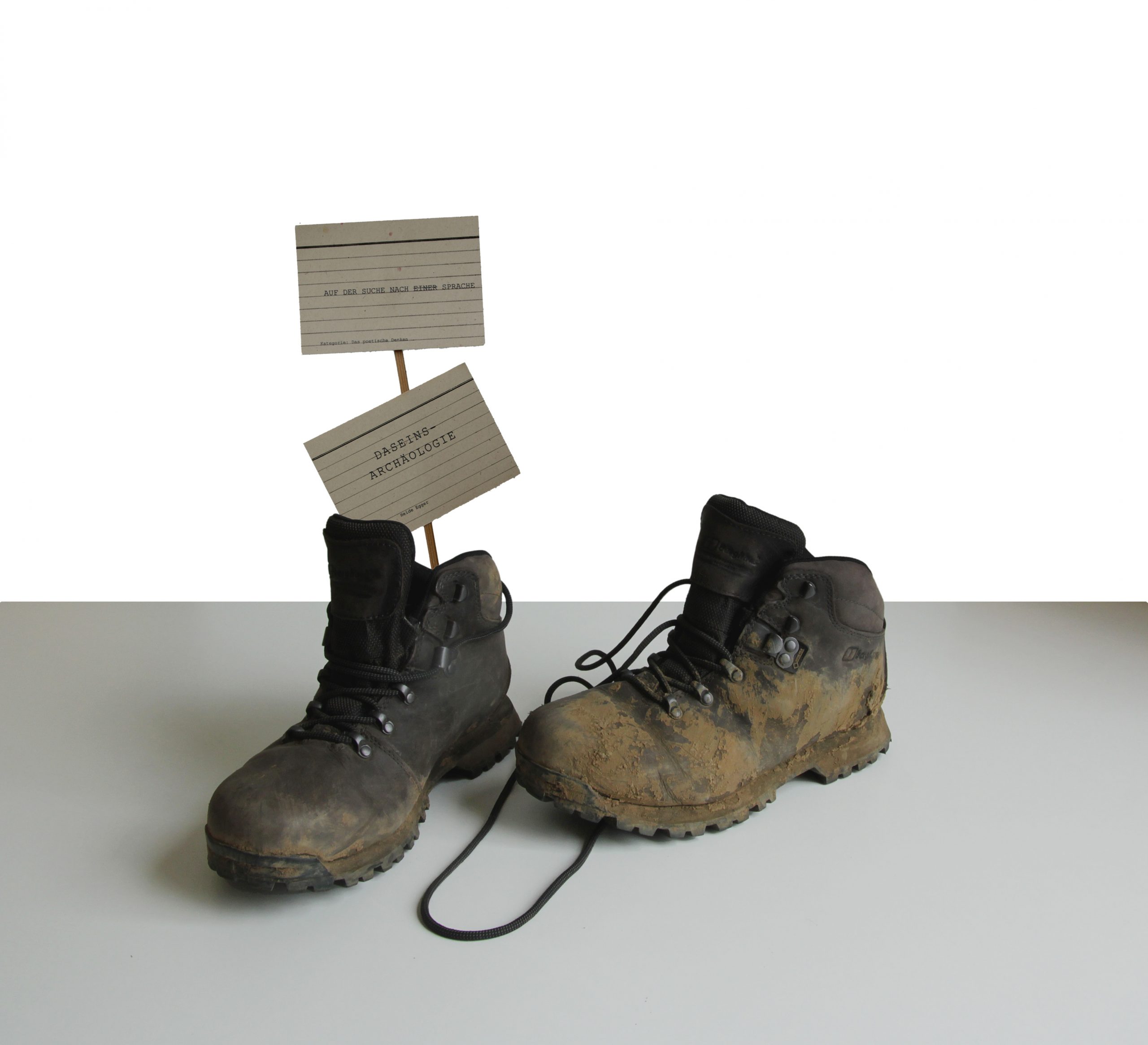 Zwei dreckige Wanderschuhe, wobei im linken Schuh zwei bedruckte Karteikarten auf Holzstielen stecken.