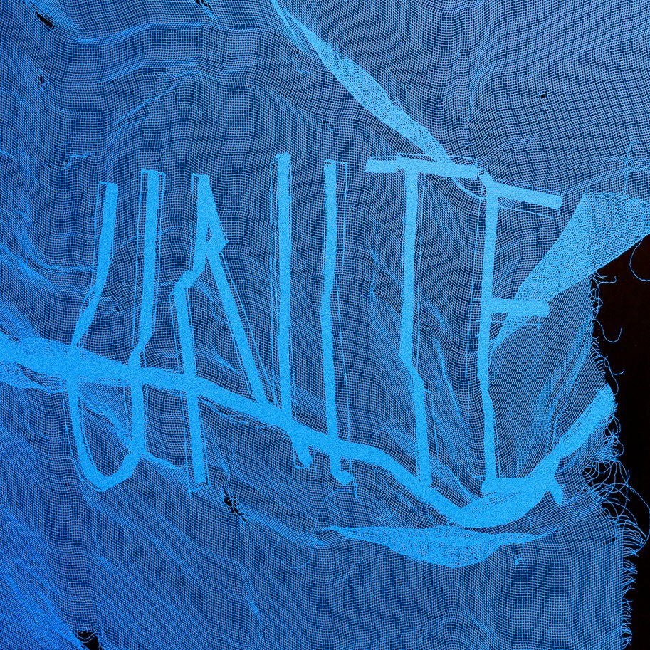 Auf schwarzem Hintergrund liegt ein durchlässiger, strahlend blauer Stoff. Auf dem Stoff ist das eingenähte Wort "Unite" zu lesen.