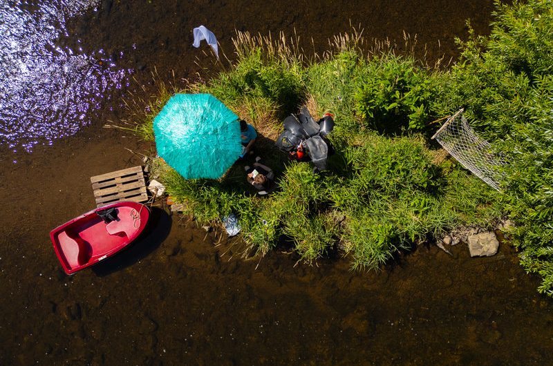 Luftaufnahme einer kleinen begrünten Insel in einem Fluss. Auf der Insel stehen zwei Personen unter einem türkisfarbenen Sonnenschirm, daneben liegt ein rotes Ruderboot und ein Holzpalette dient als Steg.