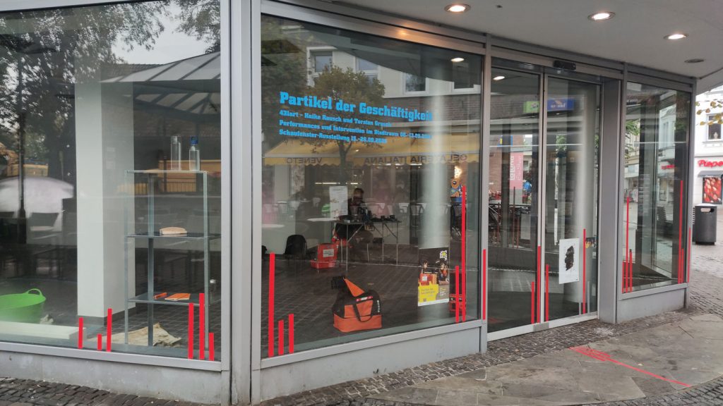 Ein Schaufenster mit der Aufschrift "Partikel der Geschäftigkeit". Innen sind undeutlich Personen zu erkennen.