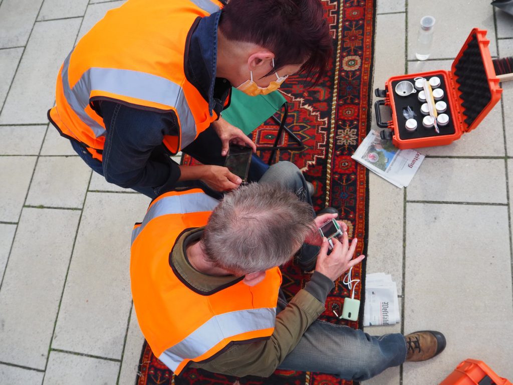 Zwei Personen mit Warnweste aus der Vogelperspektive. Sie sitzen auf einem schmalen Teppich und haben wissenschaftliche Messgeräte zur Untersuchung in der Hand.