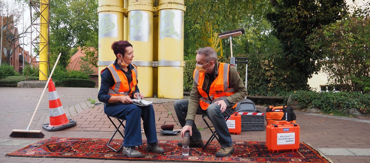 Zwei Personen in Warnweste sitzen auf Klappstühlen, die auf einem Teppich stehen, von einer Industrieanlage.
