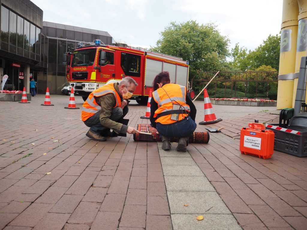 Zwei Personen mit Warnweste kehren Staub unter einen Teppich. Im Hintergrund ist ein Feuerwehrwagen zu sehen.