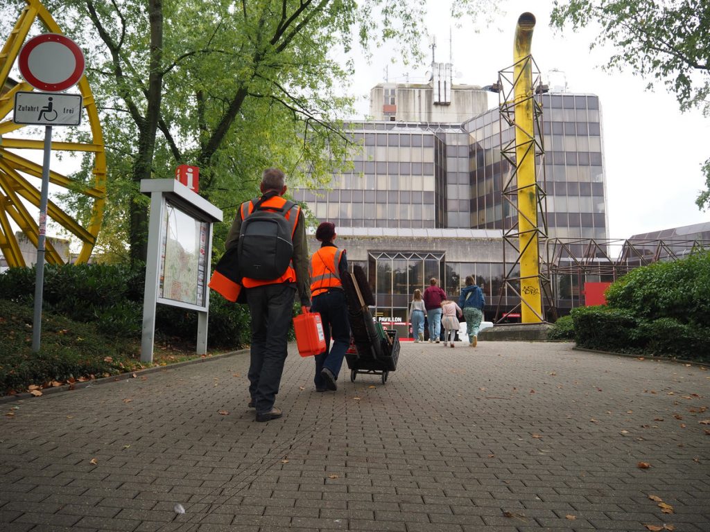 Zwei Personen mit Warnweste und neon-orangenen Koffern laufen auf ein großes Gebäude zu.