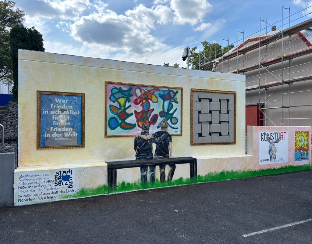 An eine Häuserfassade wurden mehrere umrahmte, kleine Bilder gemalt. Außerdem wurden zwei Personen in schwarzen T-Shirts gemalt, welche so aussehen, als würden sie die Bilder betrachten. Die gemalten Personen sichten Arm in Arm auf einer Bank.