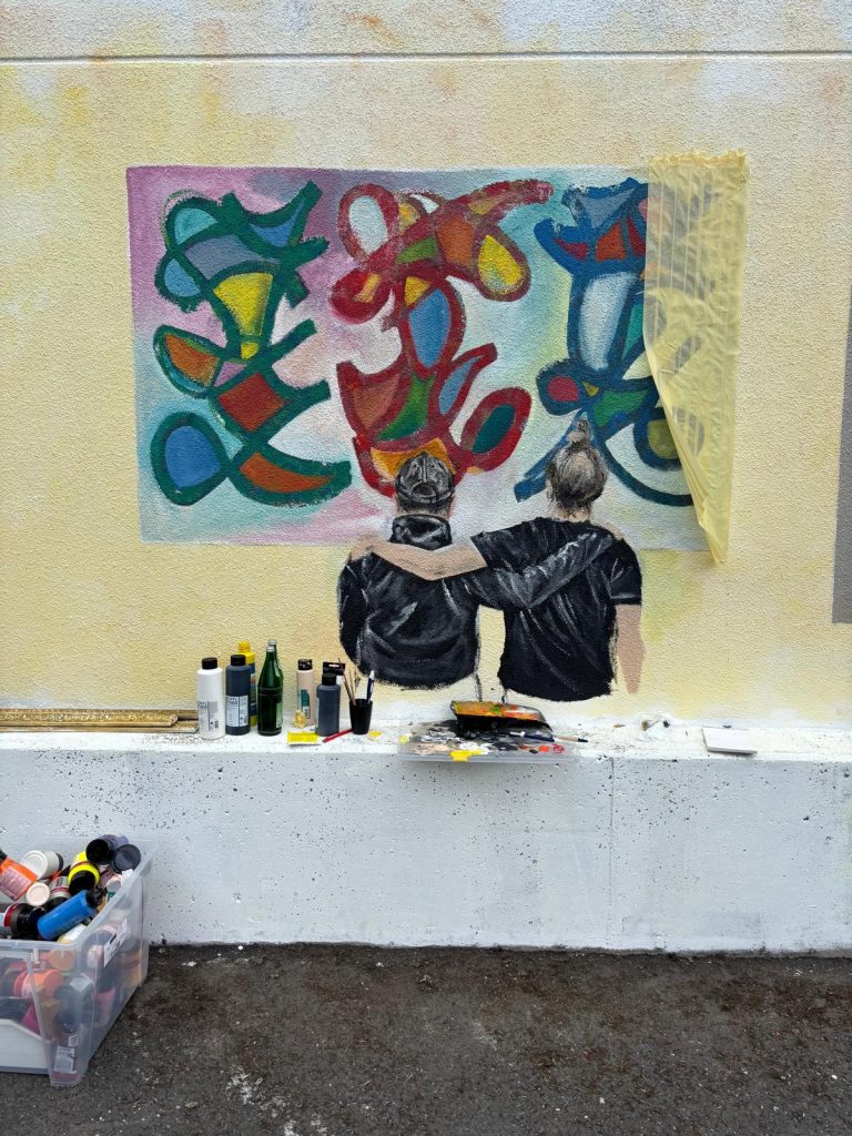 An eine Häuserfassade wurde ein Bild mit gelbem Hintergrund gemalt. Dieses Bild zeigt zwei Personen, die Arm in Arm vor einem Kunstwerk stehen und dieses betrachten. Das Kunstwerk, welches die Figuren betrachten, zeigt verschiedene Symbole in bunten Farben.