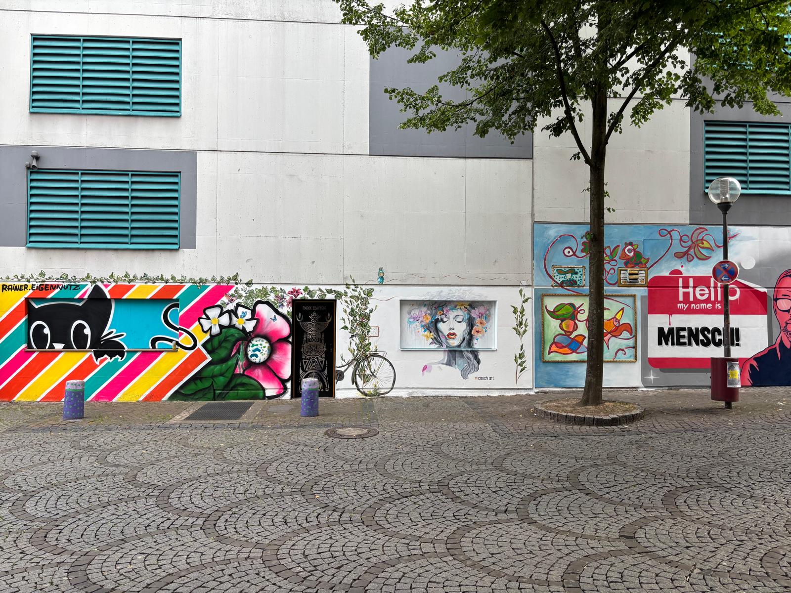 An eine graue Häuserfassade wurden viele bunte Kunstwerke gemalt. Darunter das Portrait einer Frau, ein Fahrrad, Pflanzen oder eine bunte Collage mit einer schwarzen Katze. Die Stile der Kunstwerke unterscheiden sich voneinander.