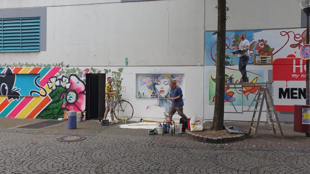 Drei Männer stehen an einer Häuserfassade und bemalen diese mit bunten Bildern. Zwei der Männer schauen in die Kamera, der dritte geht und hat den Blick nach unten gerichtet.
