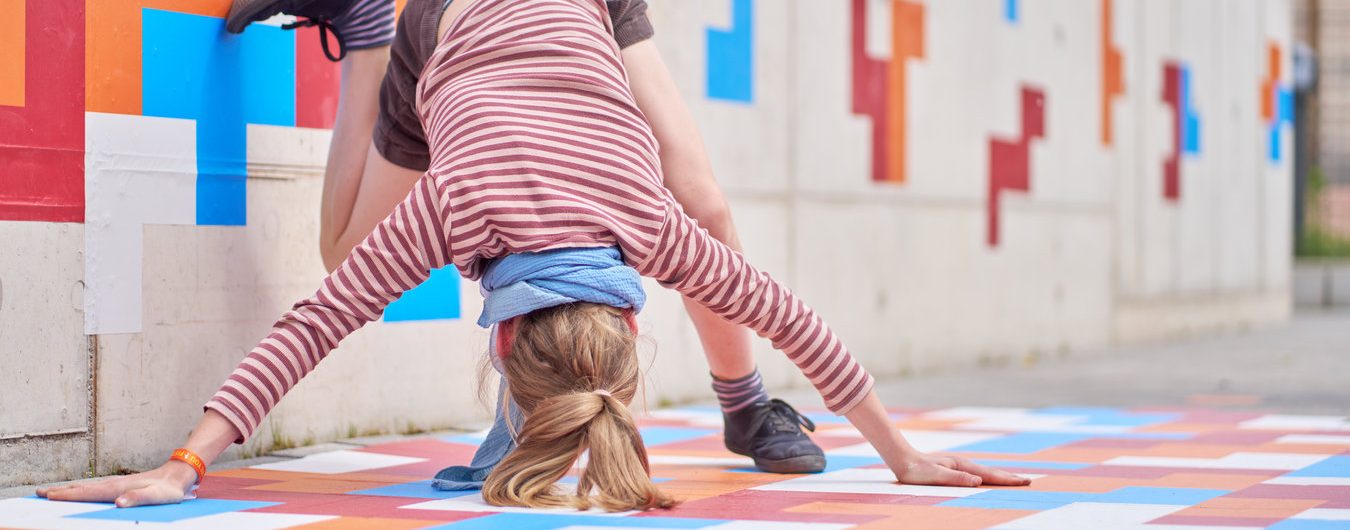 Eine junge Person ist im Beginn, einen Kopfstand auf einem mit bunten geometrischen Stickern beklebten Boden zu machen.