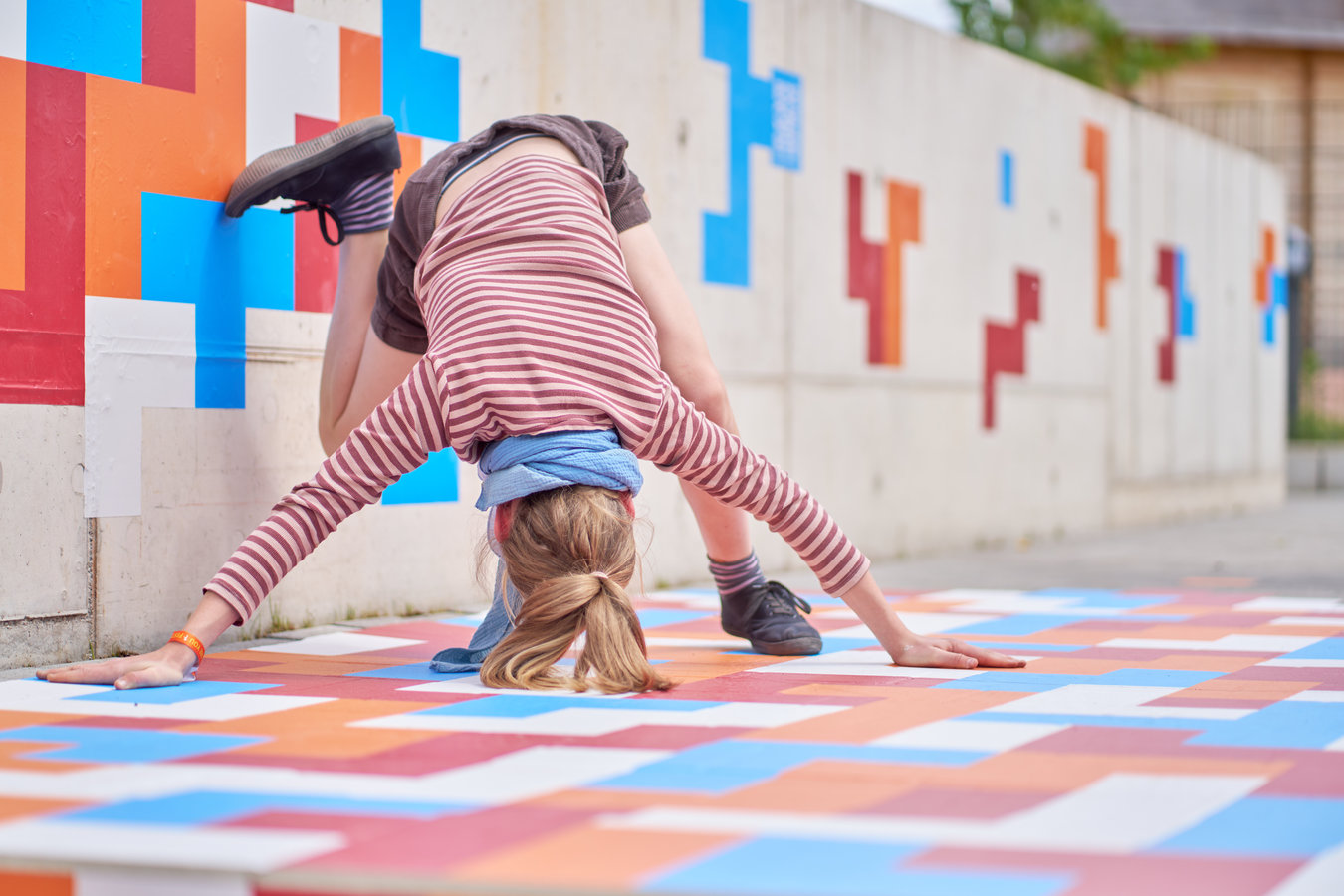 Eine junge Person ist im Beginn, einen Kopfstand auf einem mit bunten geometrischen Stickern beklebten Boden zu machen.