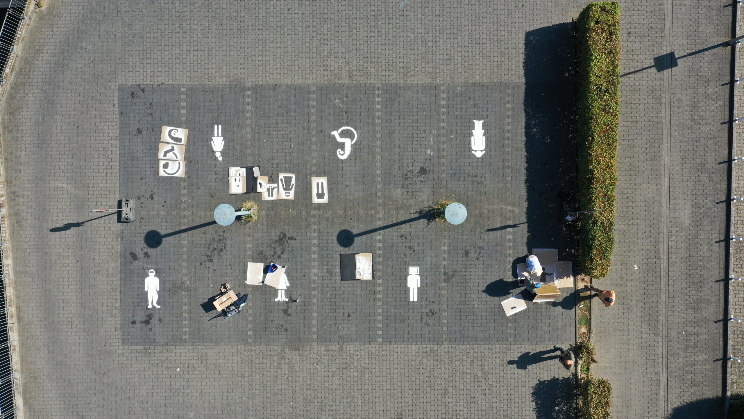 Luftaufnahme eines Parkplatzes mit mehreren markierten Stellplätzen, auf denen anstelle von Autos Piktogramme verschiedener menschlicher Figuren aufgemalt sind. Um die Markierungen herum liegen Schablonen und Malutensilien, einige Personen sind gerade dabei, weitere Figuren aufzumalen. Die Szene spielt auf einem gepflasterten Parkplatz mit zwei Laternen und angrenzender Hecke.