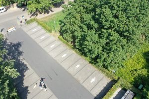 Luftaufnahme eines Parkplatzes an einer von Bäumen gesäumten Straße. Auf jedem Stellplatz ist eine unterschiedliche menschliche Figur als Piktogramm aufgemalt. Drei Personen stehen auf dem unteren Teil des Parkplatzes, daneben ein Hund. Die Szene spielt bei sonnigem Wetter und zeigt einen kreativ gestalteten öffentlichen Raum.