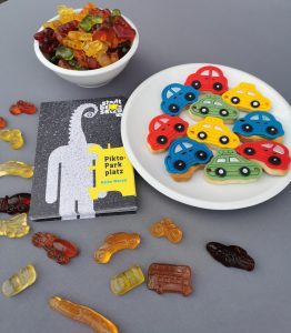 Nahaufnahme eines Tisches mit bunten Autokeksen auf einem Teller, einer Schale mit Gummibärchen und einem kleinen Heft mit dem Titel „Pikto-Parkplatz“ von Käthe Wenzel. Rund um das Heft sind Gummibärchen in Form von Fahrzeugen wie Fahrrädern, Bussen und Autos verteilt. Die Szene wirkt verspielt und farbenfroh.