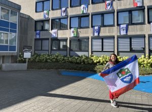 Eine Frau vor einem Verwaltungsgebäude, an dem mehrere Fahnen angebracht sind, hält selbst eine große Fahne in den Händen.