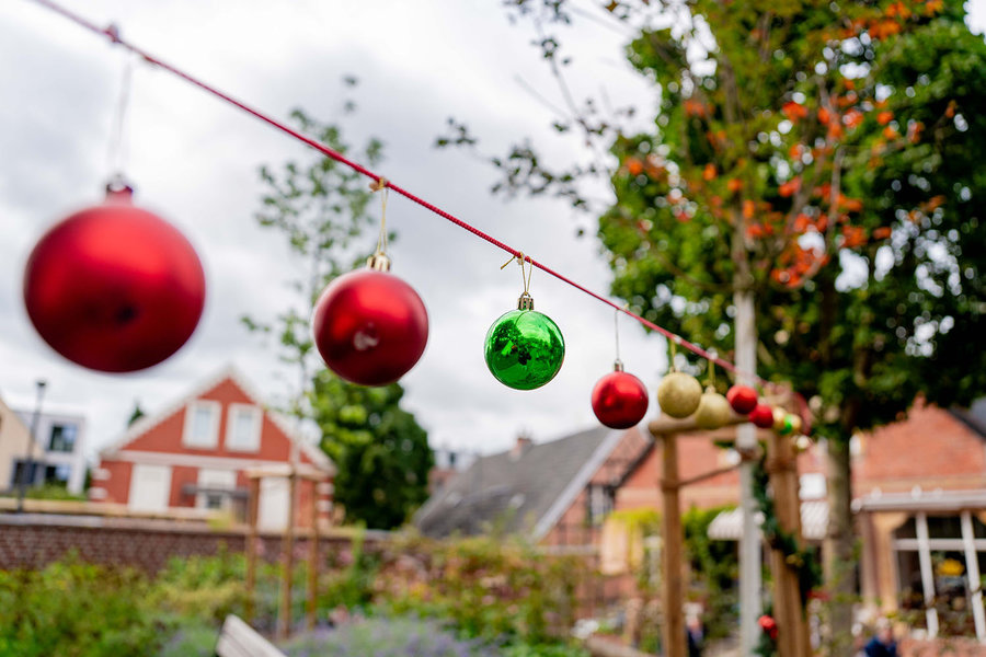 Eine Reihe bunter Weihnachtskugeln – überwiegend rot, dazu eine grüne und eine goldene – hängt an einer roten Schnur im Freien. Im Hintergrund sind Bäume mit Herbstlaub sowie einige Häuser und Gärten zu sehen. Die Aufnahme ist leicht unscharf im Hintergrund und vermittelt eine festliche, vorweihnachtliche Stimmung im Außenbereich.