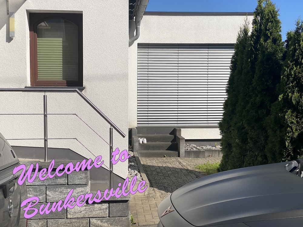 Eine Einfahrt vor einem Einfamilienhaus. In rosa Schrift steht geschrieben "Willkommen in Bunkersville".
