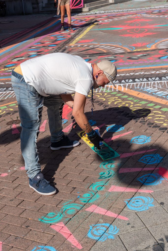 Ein Mann mit zerrissener Jeans und Kappe beugt sich nach vorne, um mit Sprühdosen bunte Muster auf einen Platz zu sprühen.