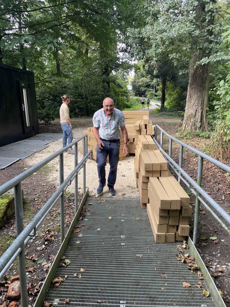 Mehrere Pappschachteln liegen aufgestapelt auf einem Weg, daneben gehen zwei Personen.