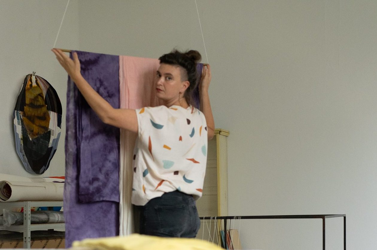 Eine Frau mit gepunktetem Oberteil hält einen Teppich in ihren Händen. Sie steht mit dem Rücken zur Kamera und schaut über ihre Schulter.