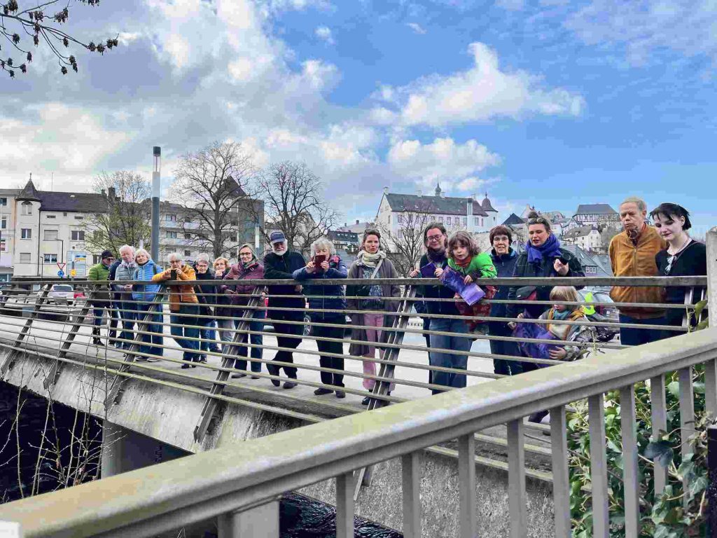 Eine Gruppe von Menschen verschiedenen Alters steht an einem Brücken-Geländer. Sie schauen alle in die gleiche Richtung und betrachten etwas. Was sie anschauen ist nicht zu erkennen.
