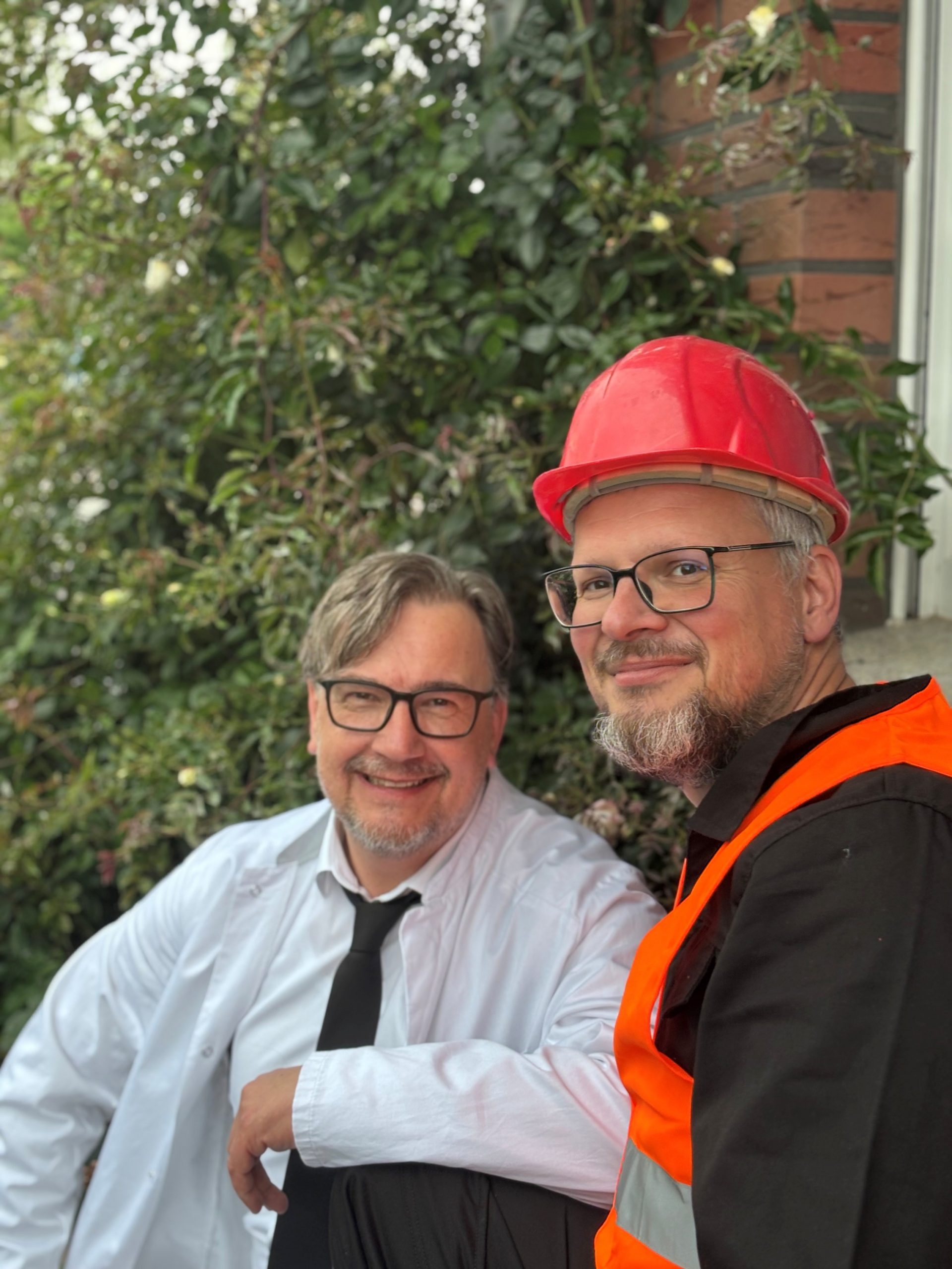 Zwei Männer sitzen draußen vor einer begrünten Hauswand und schauen lächelnd in die Kamera. Der Mann rechts trägt eine orangefarbene Warnweste und einen roten Schutzhelm, der Mann links ein weißes Hemd mit schwarzer Krawatte. Beide tragen Brillen und haben graumeliertes Haar sowie Bart. Die Atmosphäre wirkt entspannt und freundlich.