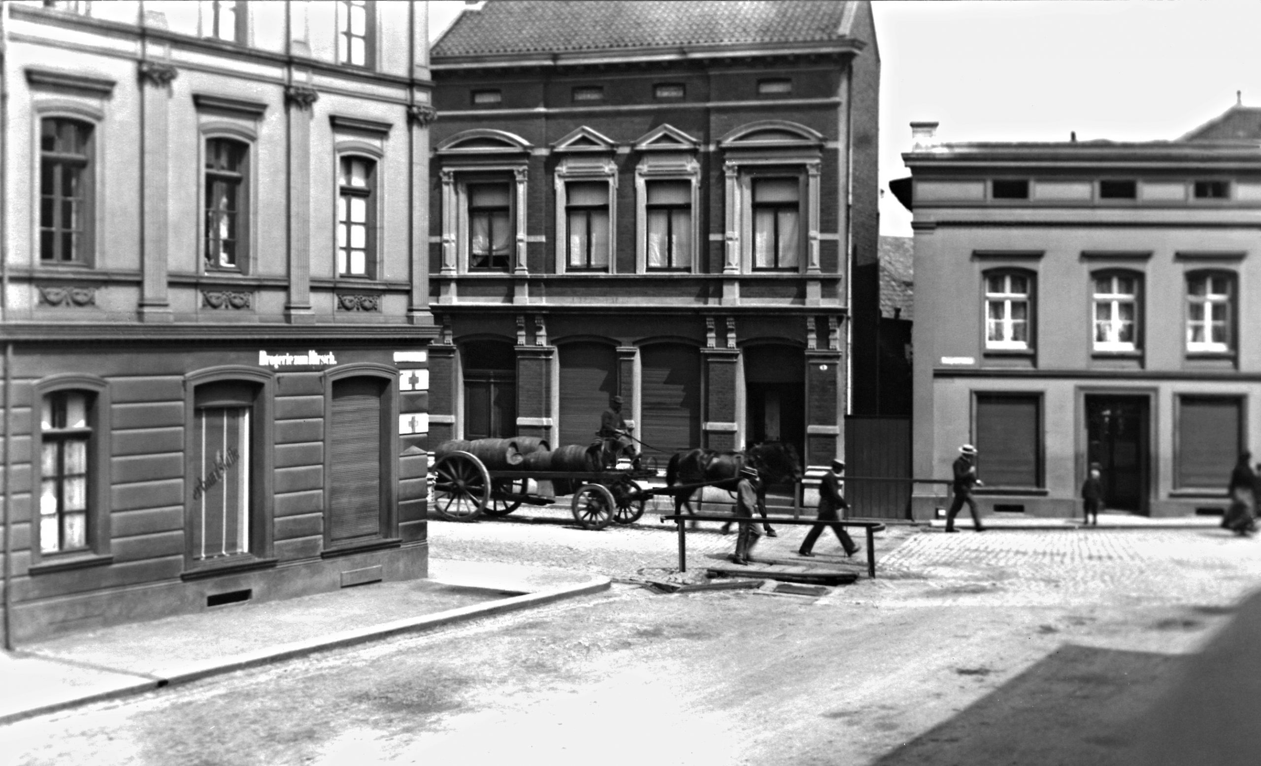 Historische Schwarz-Weiß-Aufnahme einer Straßenkreuzung mit mehrstöckigen Gebäuden aus der Gründerzeit. Auf dem Kopfsteinpflaster fährt ein von Pferden gezogener Wagen mit Fässern, mehrere Menschen überqueren die Straße zu Fuß. Im Vordergrund verläuft eine kleine Brücke oder Überführung mit Geländer. Die Szene vermittelt den Eindruck eines städtischen Alltags Anfang des 20. Jahrhunderts.