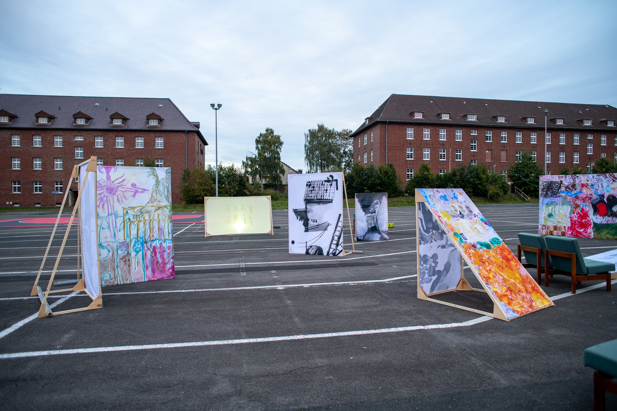 Mehrere großformatige Kunstwerke und bemalte Leinwände stehen auf einem offenen Parkplatz zwischen roten Backsteingebäuden. Die Arbeiten sind auf Holzgestellen präsentiert und bilden eine begehbare Ausstellung im öffentlichen Raum. Einzelne Sitzmöbel ergänzen die Installation unter einem bewölkten Himmel.