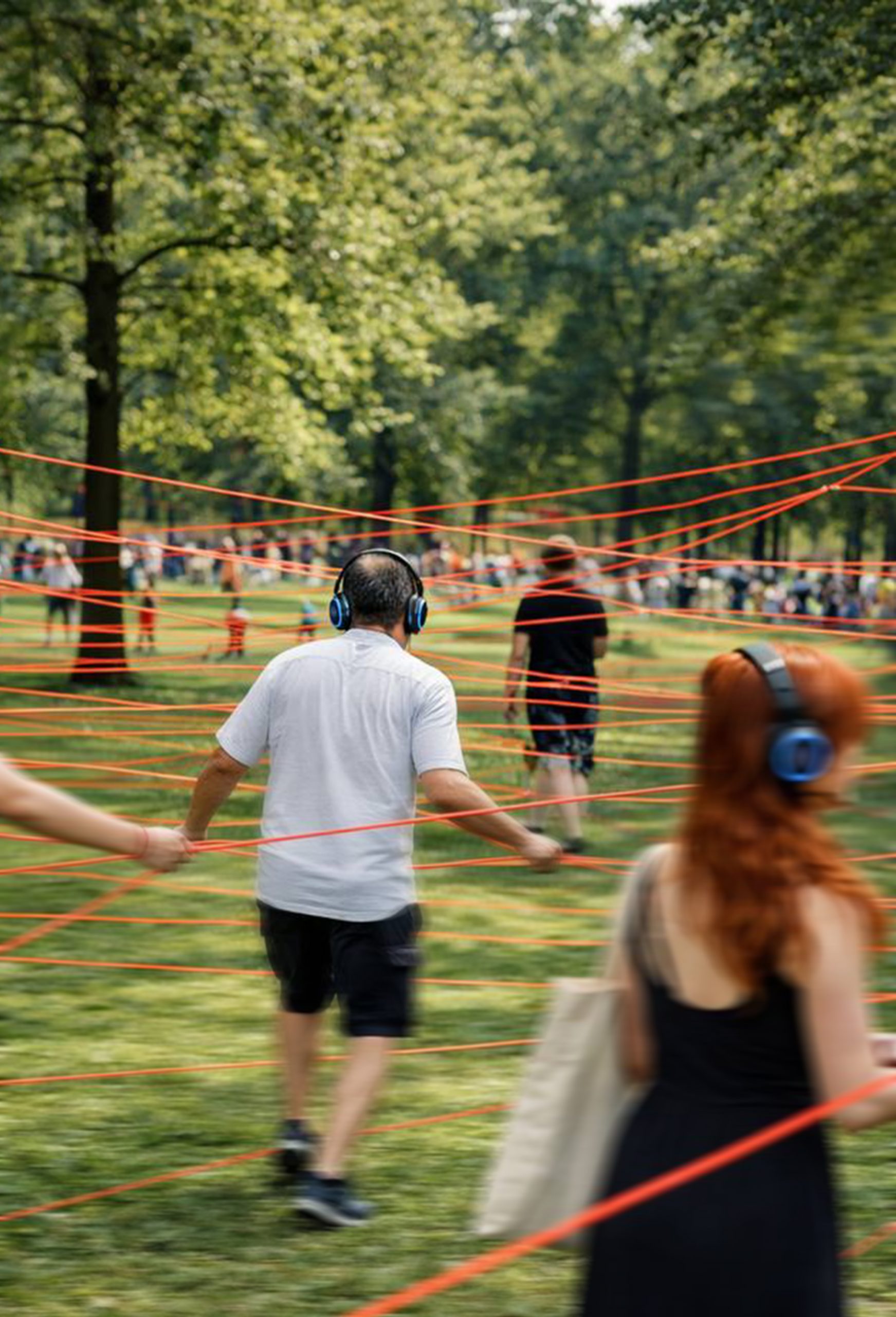 Menschen bewegen sich mit Kopfhörern durch einen Park, in dem zahlreiche rote Schnüre zwischen den Bäumen gespannt sind. Im Vordergrund geht eine Person vorsichtig zwischen den Linien hindurch, während weitere Besucher:innen im Hintergrund Teil der begehbaren Installation sind. Die sommerliche Szene vermittelt Bewegung, Orientierung und gemeinschaftliches Erleben im öffentlichen Raum.
