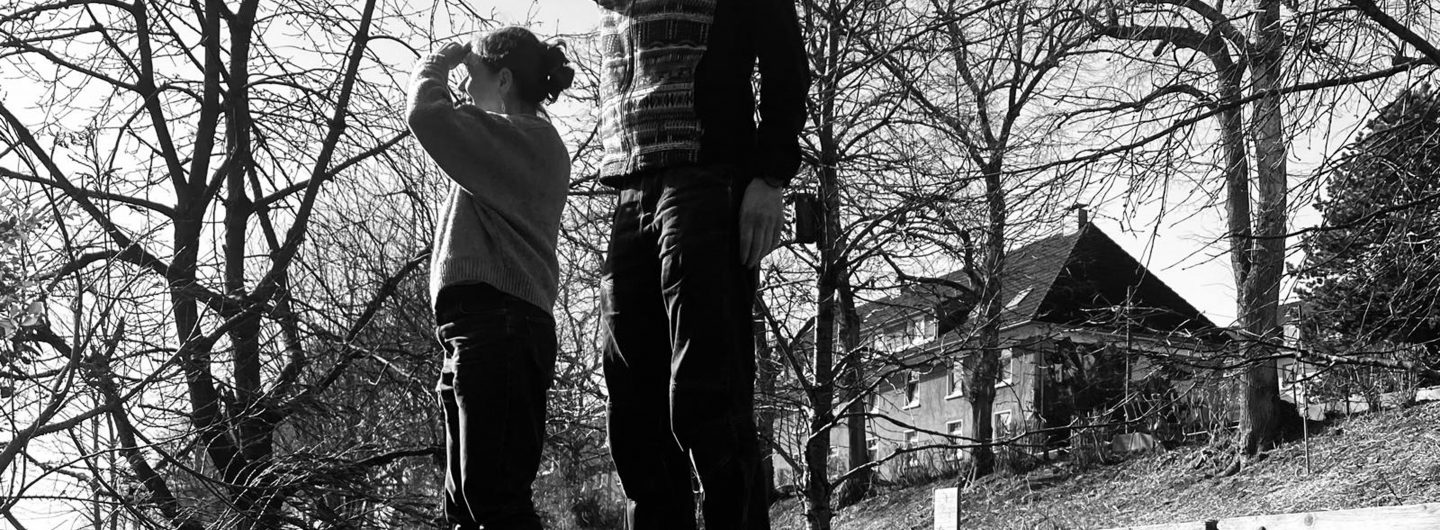 Schwarzweißfotografie von zwei Personen, die auf einem großen gefällten Baumstamm in einer winterlichen Landschaft stehen. Beide halten die Hand schützend über die Augen und blicken in die Ferne. Um sie herum stehen kahle Bäume; im Hintergrund sind Häuser an einem Hang zu sehen. Die Perspektive von unten lässt die Personen groß und suchend wirken.