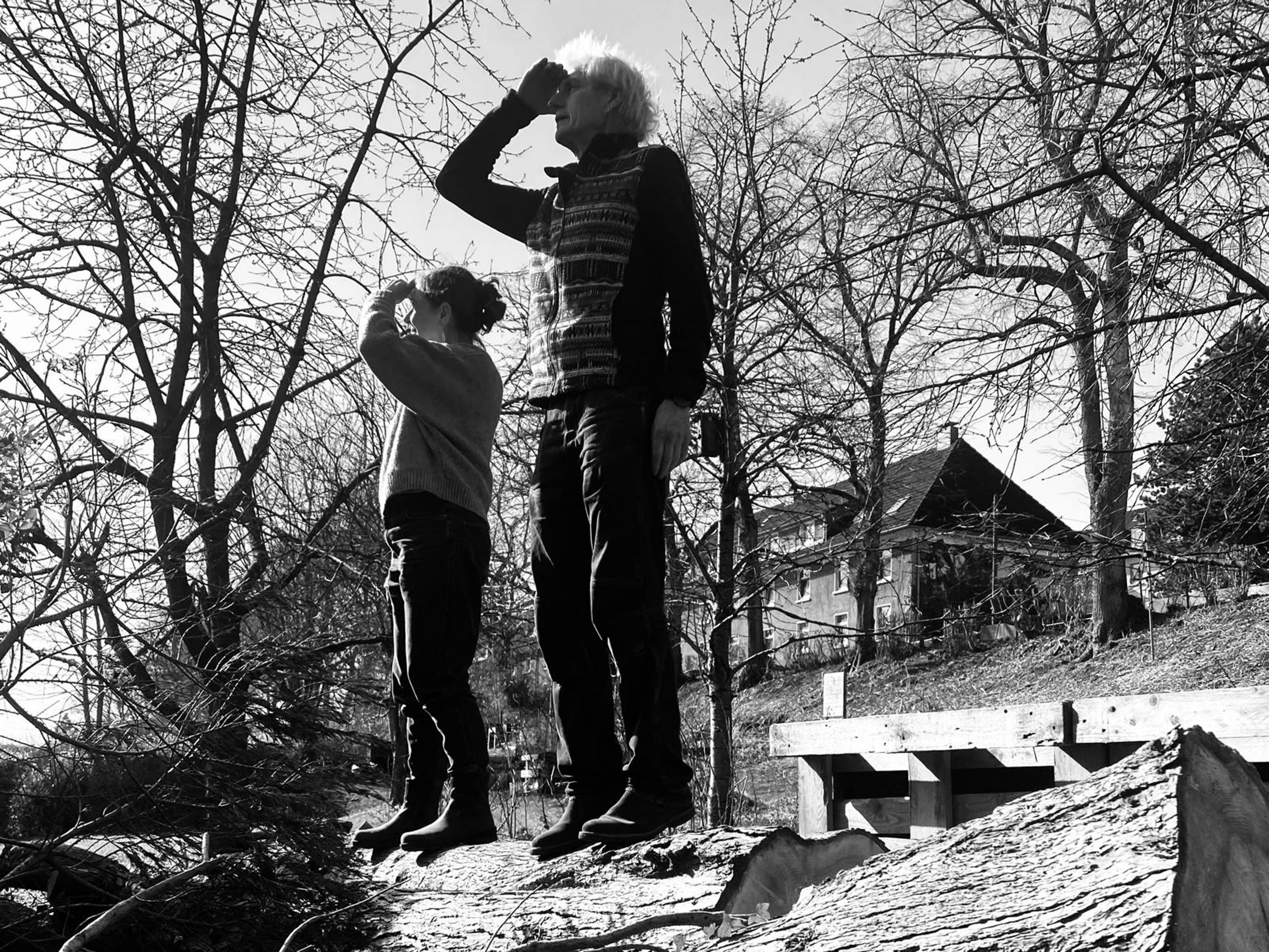 Schwarzweißfotografie von zwei Personen, die auf einem großen gefällten Baumstamm in einer winterlichen Landschaft stehen. Beide halten die Hand schützend über die Augen und blicken in die Ferne. Um sie herum stehen kahle Bäume; im Hintergrund sind Häuser an einem Hang zu sehen. Die Perspektive von unten lässt die Personen groß und suchend wirken.
