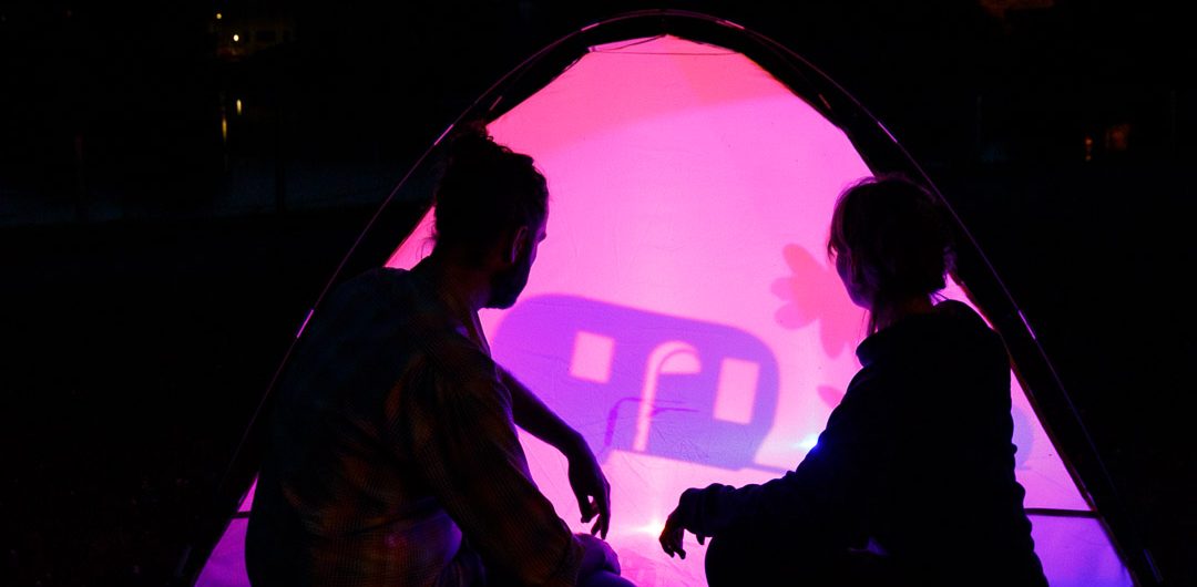 Zwei Personen sitzen in der Dämmerung vor einem von innen pink und violett beleuchteten Zelt. Auf der Zeltwand ist das Schattenspiel einer stilisierten Figur zu sehen. Im Hintergrund zeichnen sich ein See, Bäume und der dunkle Abendhimmel ab.