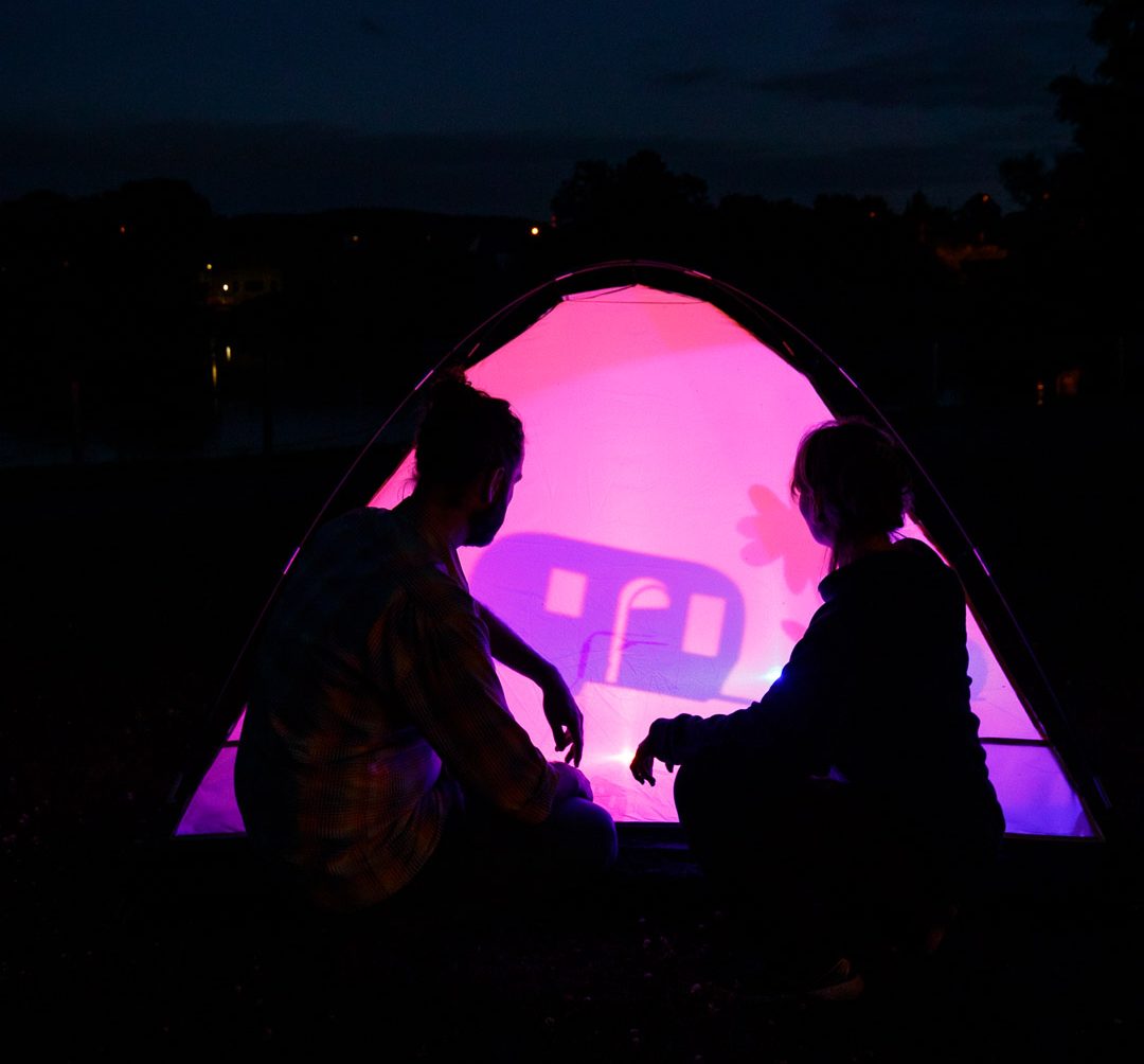 Zwei Personen sitzen in der Dämmerung vor einem von innen pink und violett beleuchteten Zelt. Auf der Zeltwand ist das Schattenspiel einer stilisierten Figur zu sehen. Im Hintergrund zeichnen sich ein See, Bäume und der dunkle Abendhimmel ab.