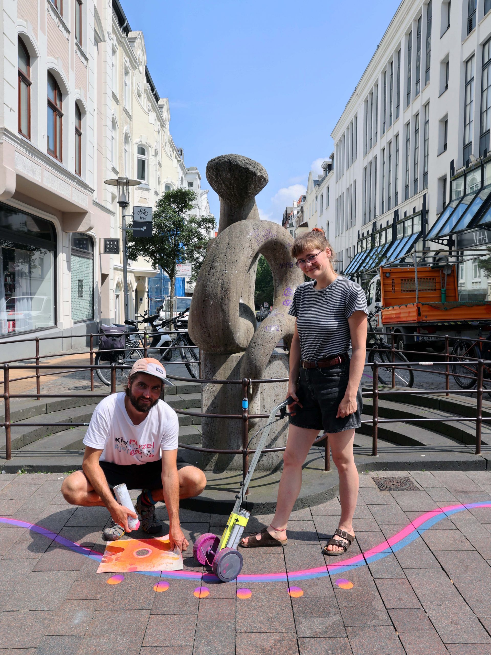 Eine junge Frau und ein junger Mann stehen vor einer Skulptur. Um sie herum wurde mit Farbe auf den Boden gemalt.