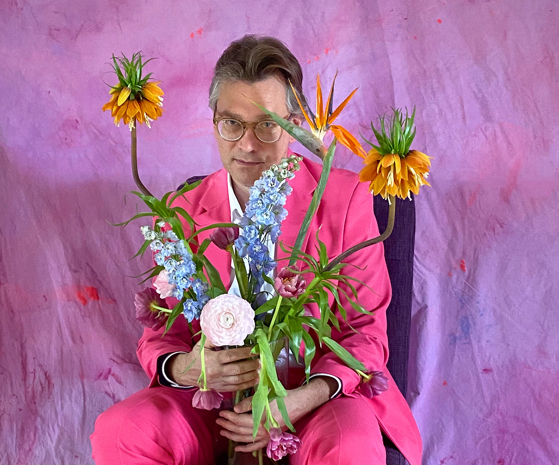 Ein Mann sitzt vor einem Lila Hintergrund. Er trägt einen pinken Anzug. In seinen Händen hält er einen Strauss Blumen.