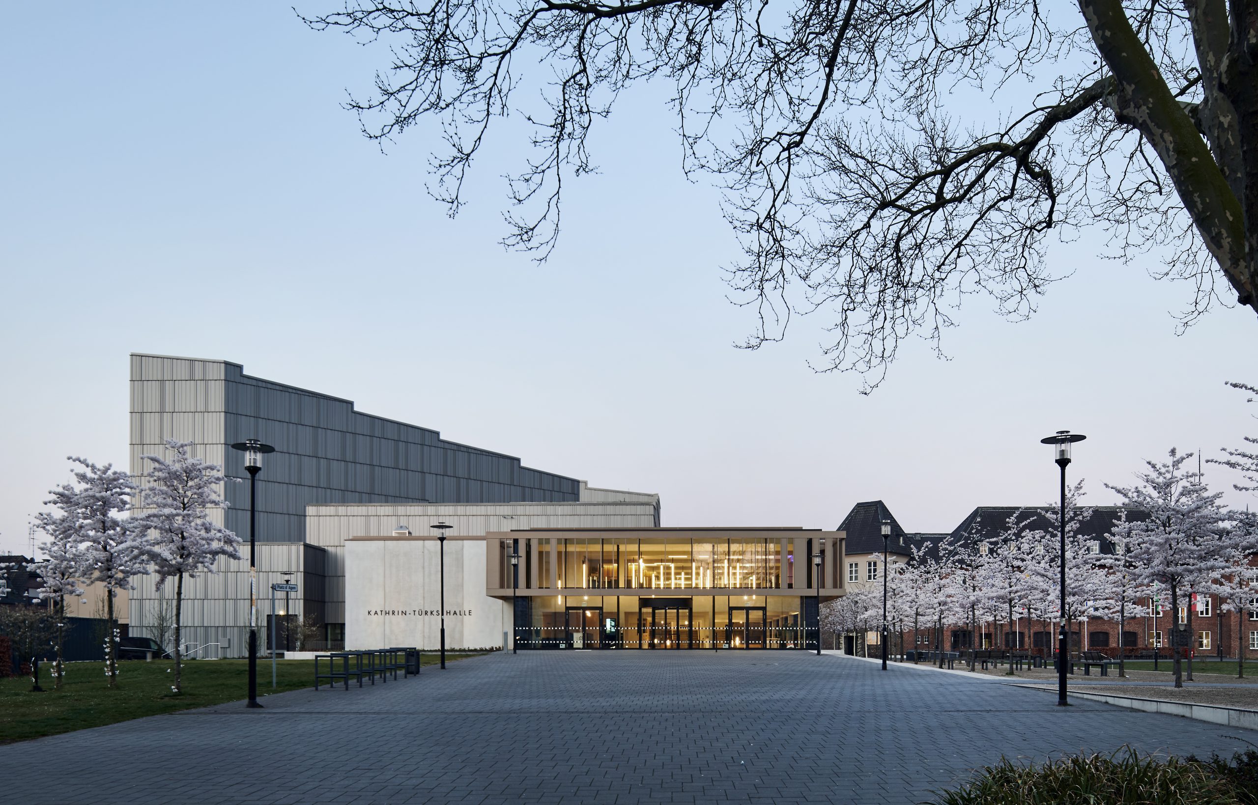 Ansicht der Kathrin-Türks-Halle mit ihrem modernen Eingangsbereich und dem angrenzenden Theatergebäude. Der Vorplatz ist leer, blühende Bäume säumen den Weg, und durch die Glasfassade fällt warmes Licht nach außen.