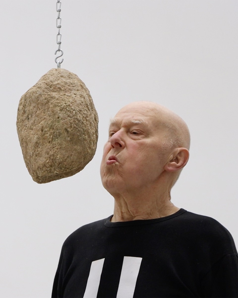 Ein älterer, kahlköpfiger Mann in einem schwarzen Oberteil mit zwei weißen Balken spitzt die Lippen in Richtung eines großen, rauen Steins, der an einer Metallkette dicht vor seinem Gesicht hängt. Der Hintergrund ist hellgrau.